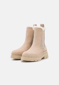 Beige ankle boots made of suede, featuring a soft fur lining at the top, elastic side panels, and a chunky rubber sole for traction.