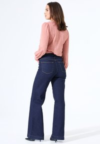 Ausgestellte dunkelblaue Jeans mit fünf Taschen, kombiniert mit einer strukturierten, rosa Bluse mit langen Ärmeln. Glattes Material und minimale Nähdetails.