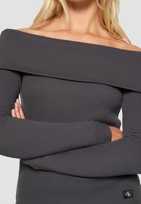 Grå ribbad långärmad topp med off-shoulder design och logotypmärke längst ner till höger. Slät textur, figurnära passform.