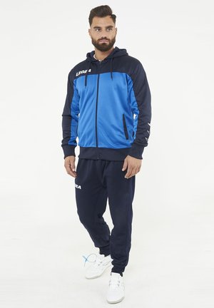 Bärtiger Mann trägt einen blau-schwarzen Kapuzenpullover mit Reißverschluss und passende schwarze Jogginghose mit weißen Sneakers, stehend auf weißem Hintergrund.