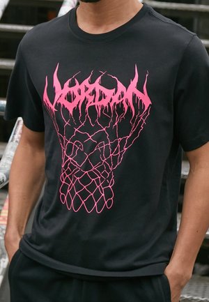 Černé tričko s růžovou grafikou připomínající basketbalovou síť a stylizovaným textem nad ní, oblečené osobou s rukama v kapsách.