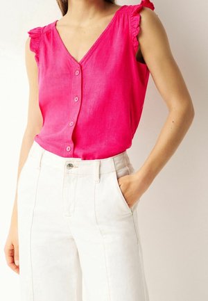 Femme portant un haut sans manches rose vif avec boutons, épaules volantées, et un pantalon blanc taille haute, une main dans la poche.