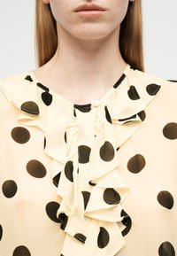 Γυναίκα που φοράει κρεμ μπλούζα με μεγάλα μαύρα πουά και γιακά με βολάν, ορατή από τον λαιμό έως το πάνω μέρος του στήθους.