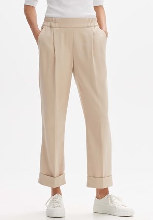 Person trägt beigefarbene, aufgekrempelte Hosen mit elastischem Bund und weiße Sneaker, die Hände in den Taschen, vor einem schlichten weißen Hintergrund.