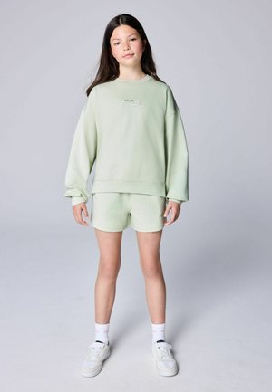 Mädchen mit langen dunklen Haaren, das ein hellgrünes Sweatshirt und passende Shorts, weiße Socken und weiße Sneakers trägt, steht vor einem einfarbigen Hintergrund.