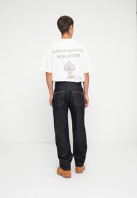 Witte t-shirt met een grijze grafische tekst en een schoppenontwerp, gecombineerd met een donkere spijkerbroek met onderscheidende hardware op de achterzakken. Bruine schoenen.