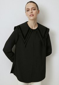 Blusa nera con un ampio colletto strutturato foderato in velluto. Maniche lunghe e spacchi laterali aumentano comfort e stile. Tessuto dal tocco morbido.