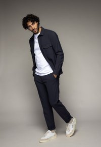 Navyblauwe knoopshirt over een witte T-shirt, gecombineerd met navy broek en witte sneakers. Glad materiaal, op maat gemaakte fit, minimalistisch ontwerp.