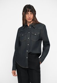 Donkerdenim shirt met lange mouwen, puntige kraag en twee borstzakken. Beschikt over contrasterend gele stiksels en zilveren drukknoopjes.