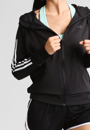 Frau trägt schwarze Adidas Zip-Jacke mit weißen Streifen an den Ärmeln über einem hellblauen Sport-BH, kombiniert mit schwarzen Shorts mit weißem Rand.