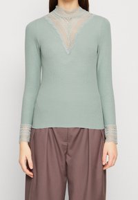 Mintgrünes Longsleeve-Oberteil mit gerippter Textur, das einen Spitzen-Ausschnitt und Spitzenakzente an den Ärmeln aufweist. Kombiniert mit lockeren braunen Hosen.