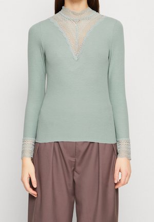 Mintgrøn langærmet bluse med ribbelsstruktur, prydet med en blondeudskæring og blondeaccenter på ærmerne, sammen med løse brune bukser.