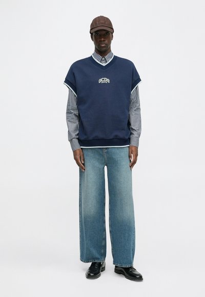 Sweatshirt sem mangas azul-marinho com logótipo, sobre uma camisa às riscas. Jeans de ganga largos e sapatos pretos completam o visual.