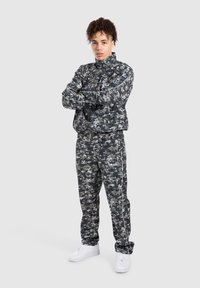 Camo-print træningsdragt i mørkegrå og sort med en lynlåsejakke og bukser med elastisk talje. Hvide detaljer og logo på bukserne.