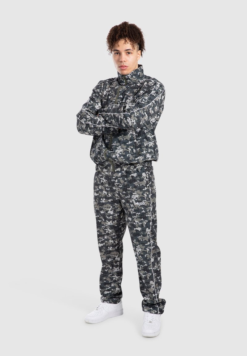 Camo-print træningsdragt i mørkegrå og sort med en lynlåsejakke og bukser med elastisk talje. Hvide detaljer og logo på bukserne.