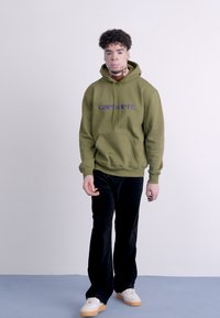 Felpa verde oliva con logo "carhartt" viola, tasca frontale, abbinata a pantaloni di velluto a coste neri e sneakers bianche con suole color gomma.
