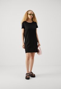 Kort svart t-shirtklänning med lös passform, texturerad tyg, rund halsringning, korta ärmar, kombinerad med kraftiga svarta sandaler och en liten rosa väska.