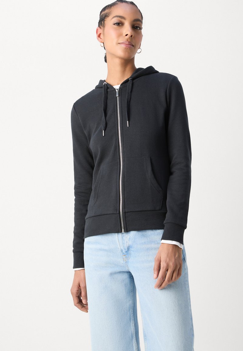 Black zip-up hoodie made of soft fabric, featuring a hood, kangaroo pockets, and silver hardware, paired with light blue straight-leg jeans.