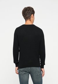 Pulover negru din tricot lână, cu gât rotund și manșete cu model în dungi, având un model texturat, purtat cu blugi albaștri.