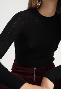 Pull en maille côtelée noire avec une coupe ajustée, une texture sophistiquée et des manches longues. Porté avec un pantalon en velours côtelé bordeaux à taille haute.