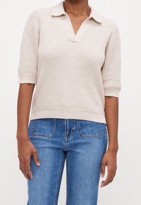 Polo en maille beige clair avec manches courtes, col en V, texture côtelée et ourlet droit, associé à un jean bleu.