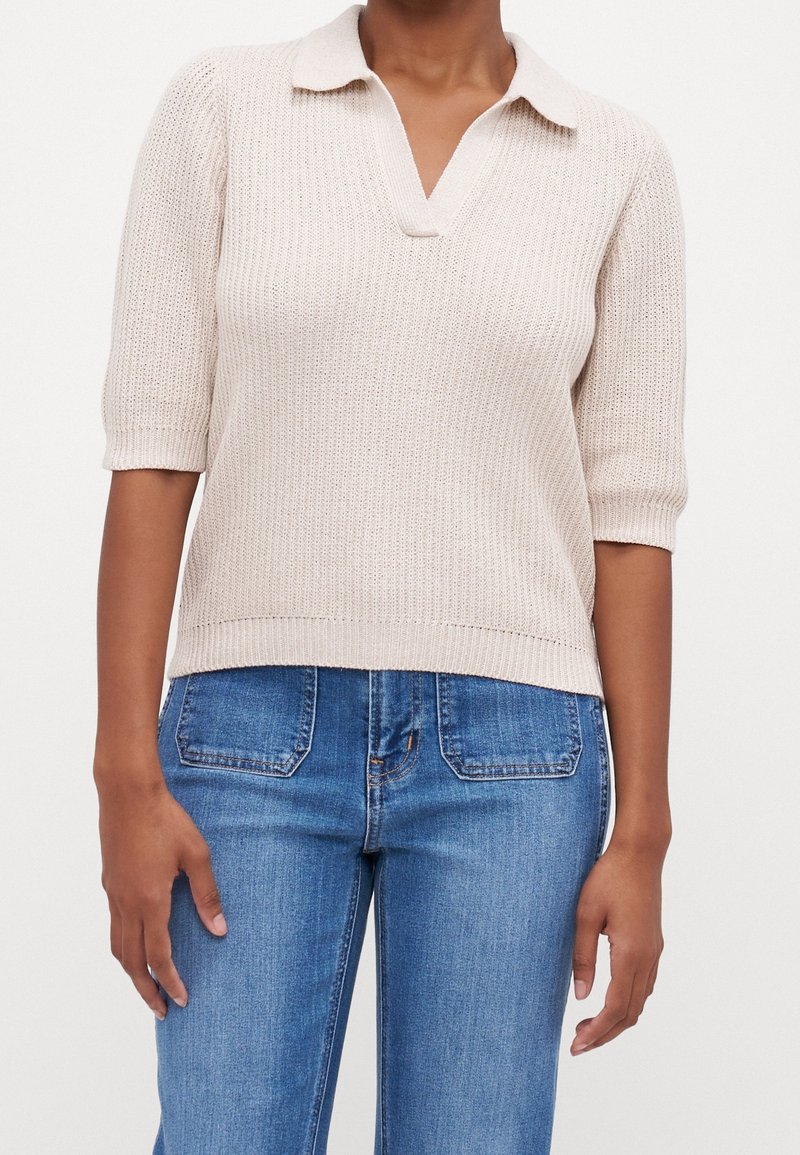 Polo en maille beige clair avec manches courtes, col en V, texture côtelée et ourlet droit, associé à un jean bleu.