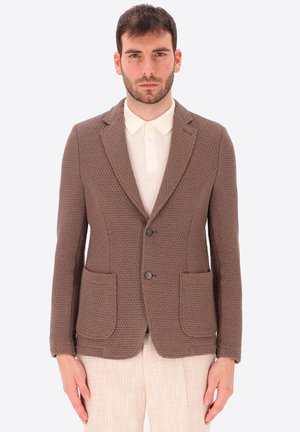 Uomo con blazer marrone testurizzato sopra una polo bianca e pantaloni beige chiaro, in piedi davanti a uno sfondo chiaro uniforme.