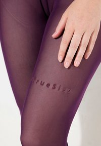 Fialové průhledné legíny s texturovaným povrchem a embosovaným logem "#TrueSize" na dolní části nohy, s rukou jemně odpočívající na látce.