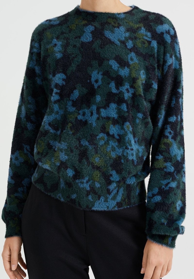 Personne portant un pull à manches longues et duveteux avec un motif floral bleu et vert, associé à un pantalon noir, debout devant un fond uni.