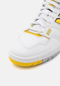 Sneaker bianco con accenti gialli, caratterizzata da una tomaia in pelle liscia, punta perforata, lacci piatti e suola in gomma texturizzata.