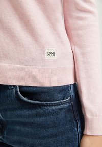 Close-up of person wearing light pink sweater with "Polo Club" label and dark blue denim jeans, showing side and lower torso.