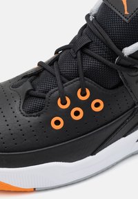 Černá atletická bota s texturovaným povrchem, oranžovými očky a síťovinovými akcenty. Obsahuje robustní tkaničky a hladkou bílou a oranžovou podrážku.