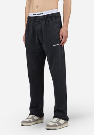 Pantalones de chándal negros con cintura elástica, logo blanco en el muslo izquierdo, bolsillos laterales y diseño de pierna recta. Combinados con zapatillas claras.