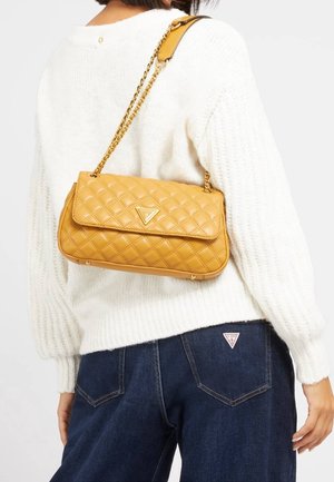 Personne portant un pull en tricot blanc et un jean foncé avec un sac à bandoulière matelassé jaune, doté d'une chaîne dorée et d'un logo triangulaire.