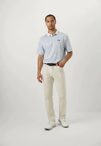 Άνδρας που στέκεται φορώντας ένα ανοιχτό μπλε πουκαμισάκι με σχέδια, μπεζ παντελόνι με μαύρη ζώνη και λευκά παπούτσια, απέναντι από έναν απλό φόντο.