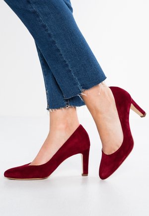 Pieds portant des chaussures en daim rouge foncé à talons moyens associées à un jean bleu court à ourlet effiloché sur un sol blanc.