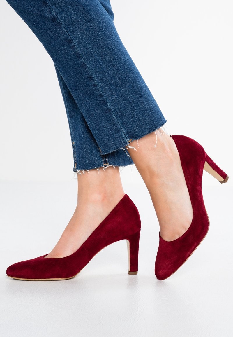 Pieds portant des chaussures en daim rouge foncé à talons moyens associées à un jean bleu court à ourlet effiloché sur un sol blanc.