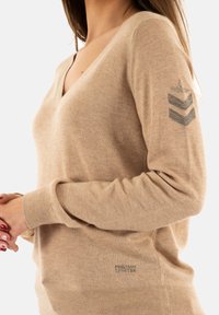 Pull en V beige en tissu doux, avec une accentuation à motifs sur la manche gauche. Bords côtelés aux poignets et à l'ourlet avec une discrète marque en bas.