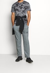 Grå camouflage t-shirt, sort sweater bundet om livet, lysegrå bukser og sorte sportsligt designede sko, alle med et afslappet sportst look.