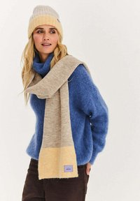 Pletený modrý svetr s vysokým límcem, béžovožlutý šál volně přehozený, s označenou štítkem. Hřejivé textury a kontrast barev zvýrazňují design.