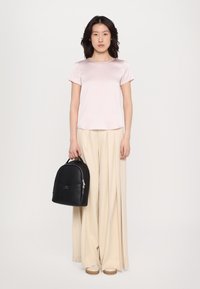 Mochila de cuero negra de forma redondeada con asa en la parte superior, combinada con una blusa de satén rosa claro y pantalones de pierna ancha plisados en beige.
