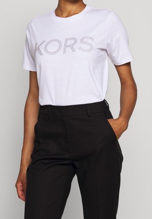 Kobieta ubrana w biały T-shirt z krótkim rękawem z napisem „KORS” z kryształków oraz czarne, wysokie spodnie z dłońmi w kieszeniach.