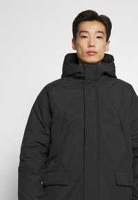 Svart pufferjakke med høy krage og hette, med to sidelommer og glidelåslukking foran. Glatt nylon tekstur.