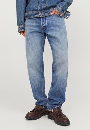 Osoba ubrana w jasnoniebieskie jeansy z denimu, kurtkę jeansową oraz ciemnobrązowe skórzane mokasyny z frędzlami, stojąca z jedną ręką w kieszeni.