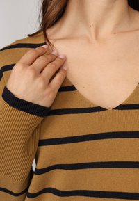 Pull rayé marron et marine avec une texture côtelée et un décolleté en V prononcé. Main visible reposant près du col.