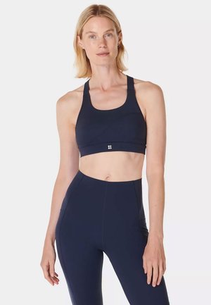 Femme aux cheveux blonds portant un soutien-gorge de sport bleu marine et un legging taille haute, debout devant un fond blanc uni.