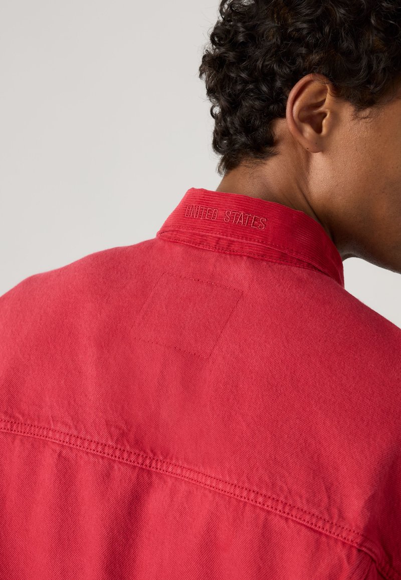 Persona con capelli ricci scuri che indossa una giacca rossa con la scritta "UNITED STATES" ricamata sul retro del colletto, mostrata da dietro e di lato.