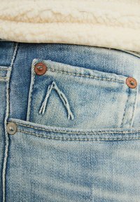 Bolsillo de vaqueros de mezclilla azul claro con un diseño triangular bordado, remaches de cobre y costuras doradas en contraste. Textura suave.