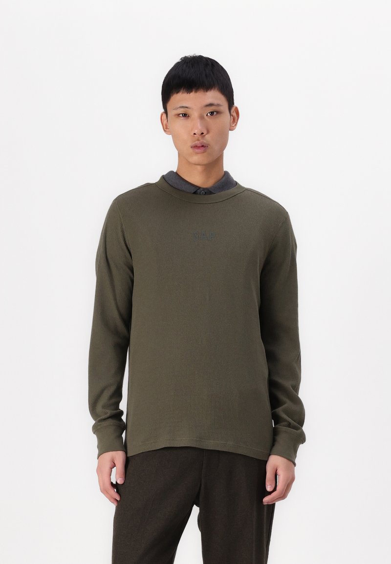 Tricou lung de culoare verde măslin, cu un model texturat, guler rotund și un detaliu subtil cu logo pe piept, asortat cu pantaloni închisi la culoare.