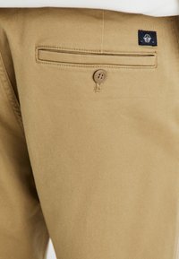 Khaki trousers made from cotton with a smooth texture. Features a back pocket with a button and a small brand label above the pocket.